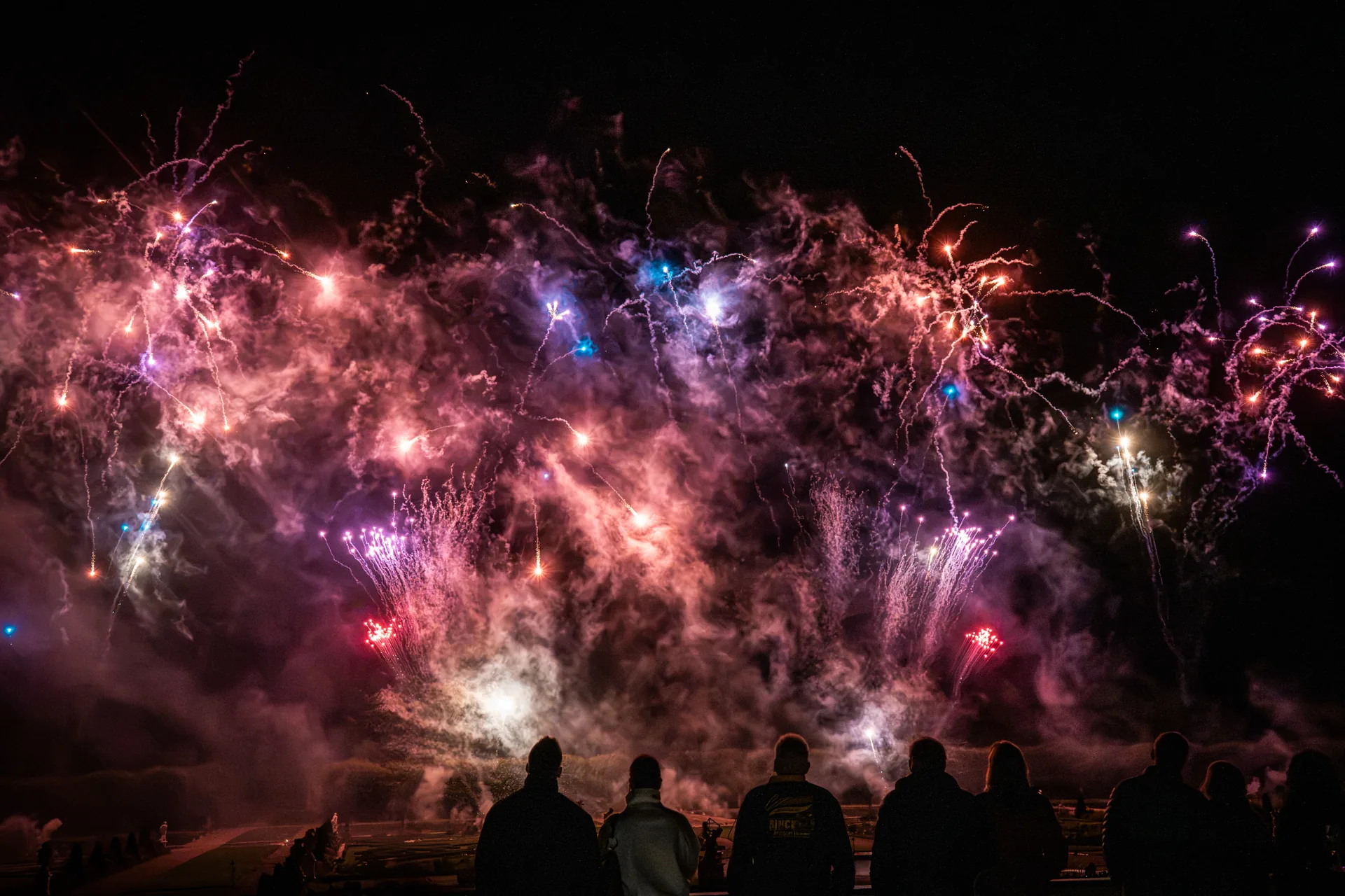 Spectators watching a wide pink and blue fireworks display